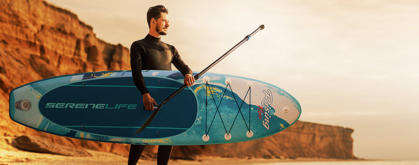 man holds a SereneLife branded paddle board on a beach with rock red cliffs behind him