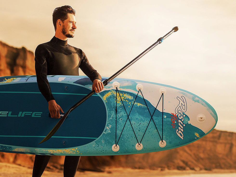 man holds a SereneLife branded paddle board on a beach with rock red cliffs behind him